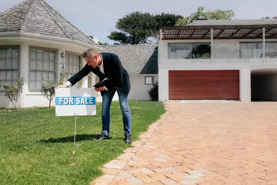 Real estate agent wearing jeans and sport coat planting For Sale sign on front lawn of home.